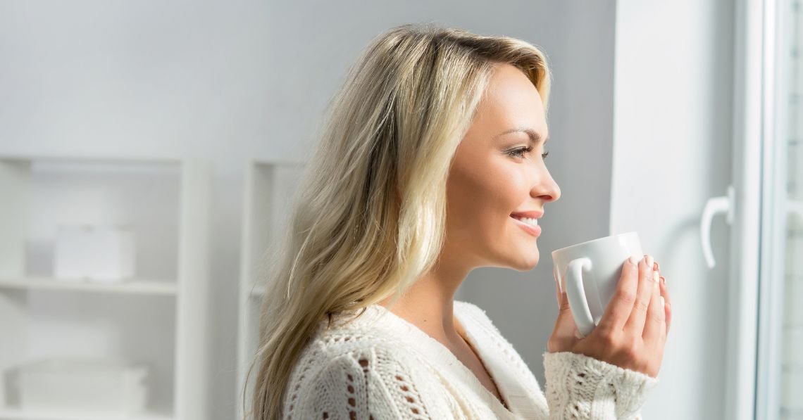 Woman in kitchen looking out a window with a cup of Live Conscious Colostrum