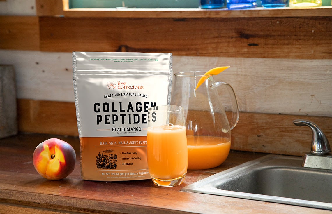 Live Conscious Peach Mango Peptides on a kitchen countertop with a peach and a glass of Peach Mango Collagen Peptides