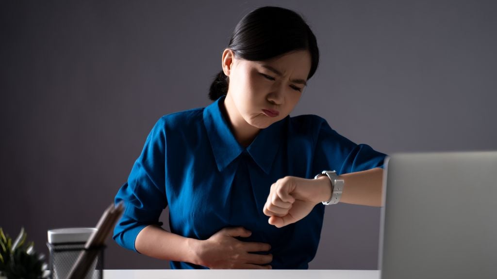 Woman at desk with one hand on stomach and the other looking at the time on her watch.