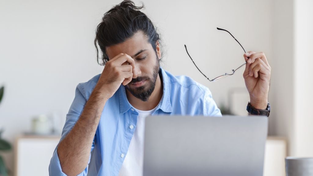 Man sitting at a computer holding glasses and feeling tired